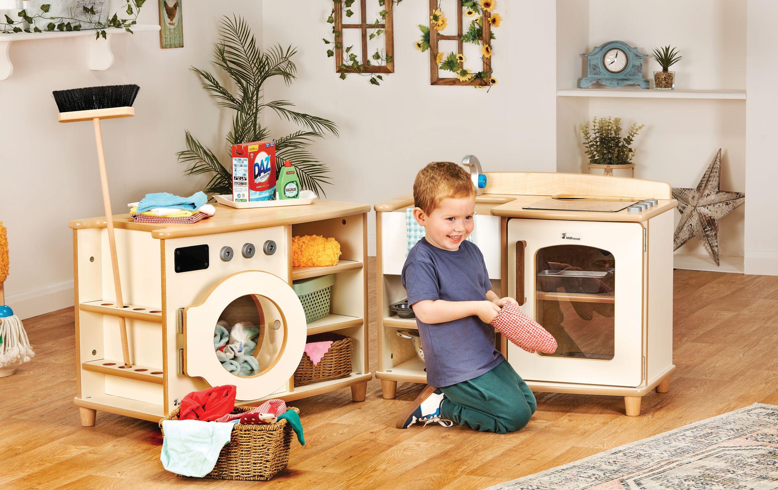 A young boy smiles while playing with a Millhouse timber play kitchen and laundry set, featuring a toy washing machine, oven, and role-play cleaning accessories in a bright nursery.