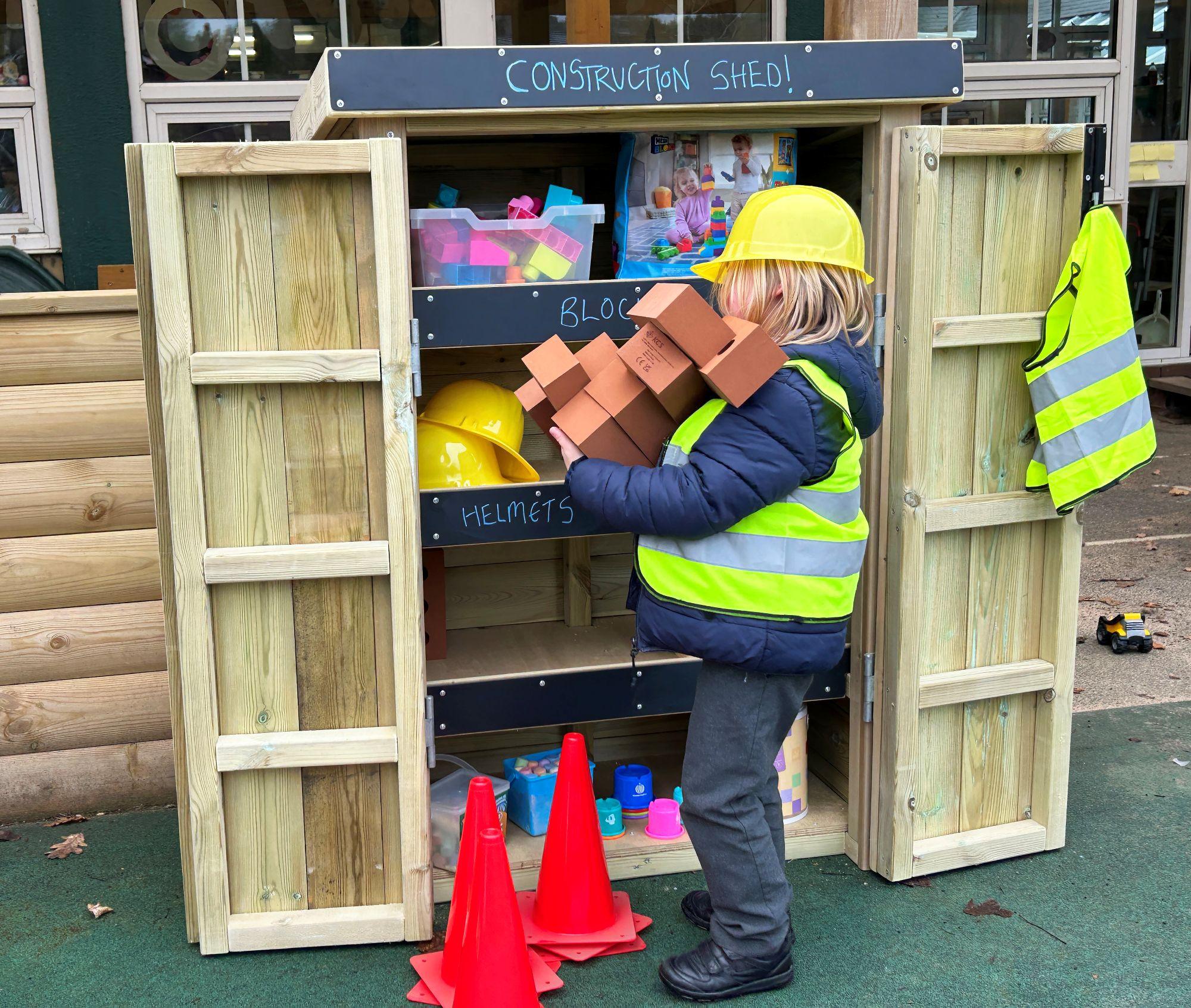 A child dressed in a hi-vis vest and yellow hard hat carries foam bricks next to a timber "Construction Shed" Acorn Store, which is filled with play helmets, blocks, and cones.