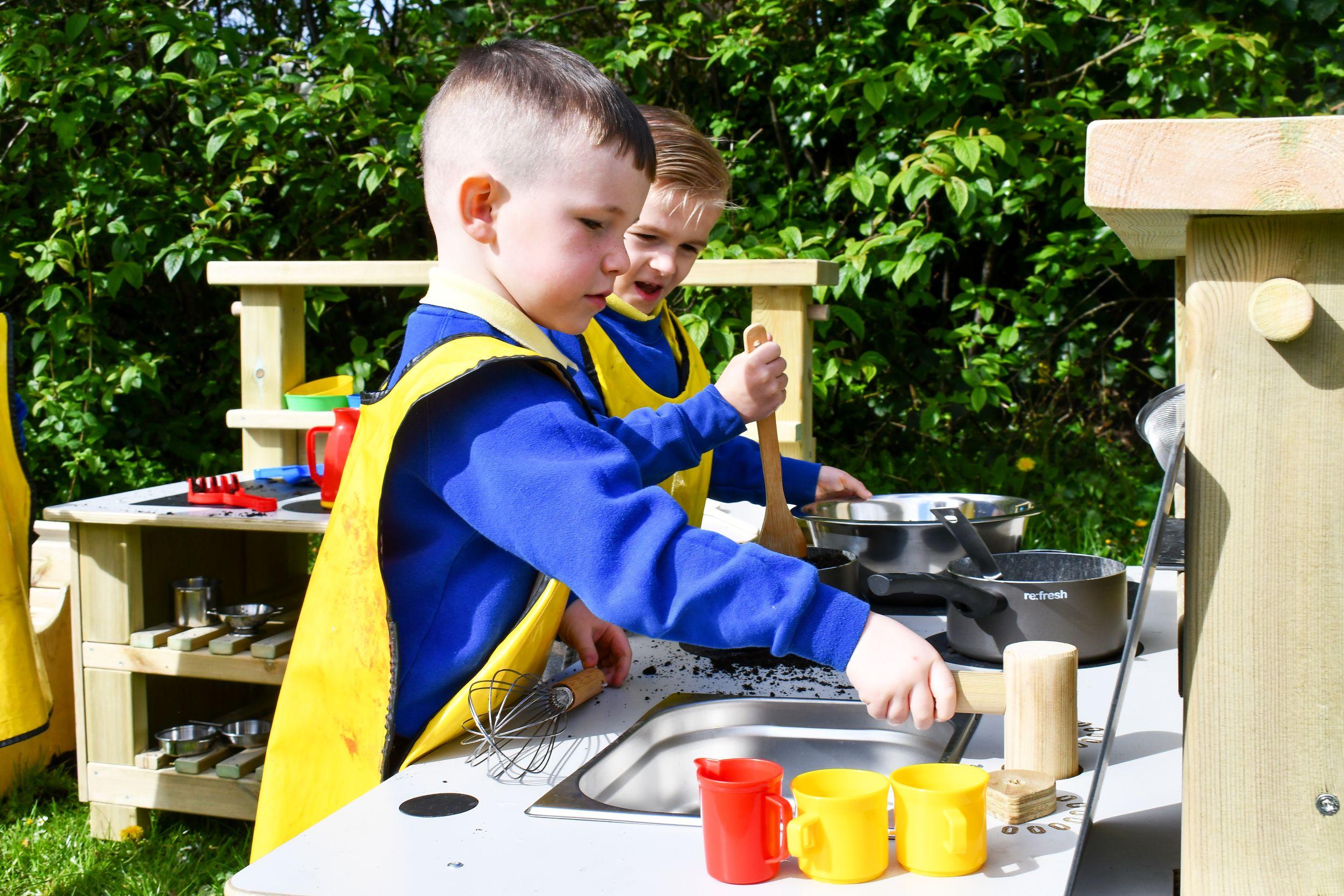 Two children in blue sweaters and yellow aprons play at a timber Mud Kitchen, using wooden spoons, whisks, and metal pots to "cook" with soil in a sunny outdoor setting.