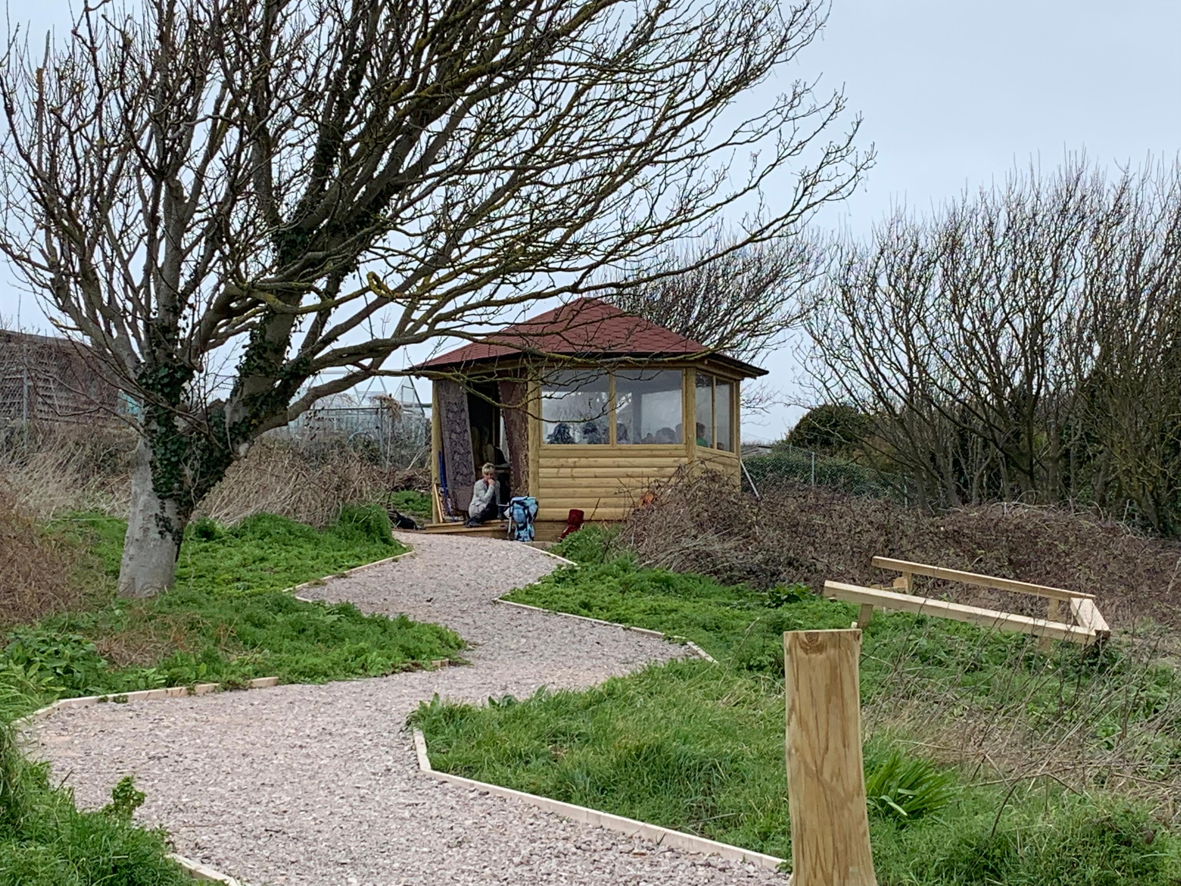 A wooden gazebo has been installed on a grass surface with a gravel path leading to the structure. A group of children can be seen inside the outdoor gazebo being taught a lesson. It is surrounded by trees and flowers.
