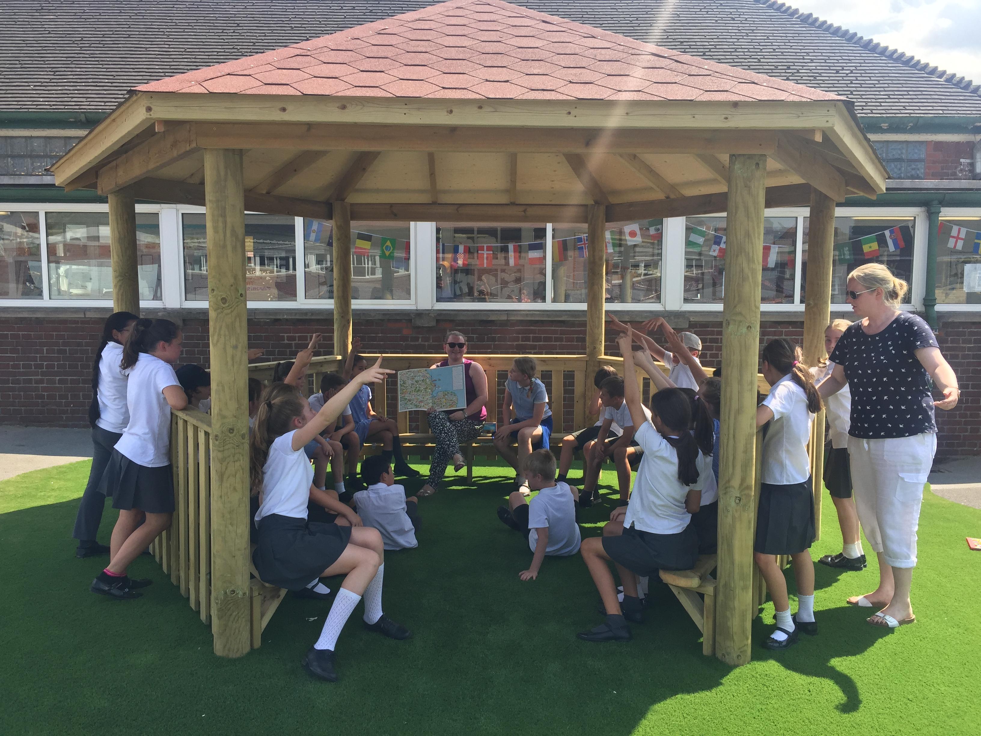 A wooden gazebo that has been installed on artificial grass with a group of children inside. A teacher is stood on the other side, looking at the children.