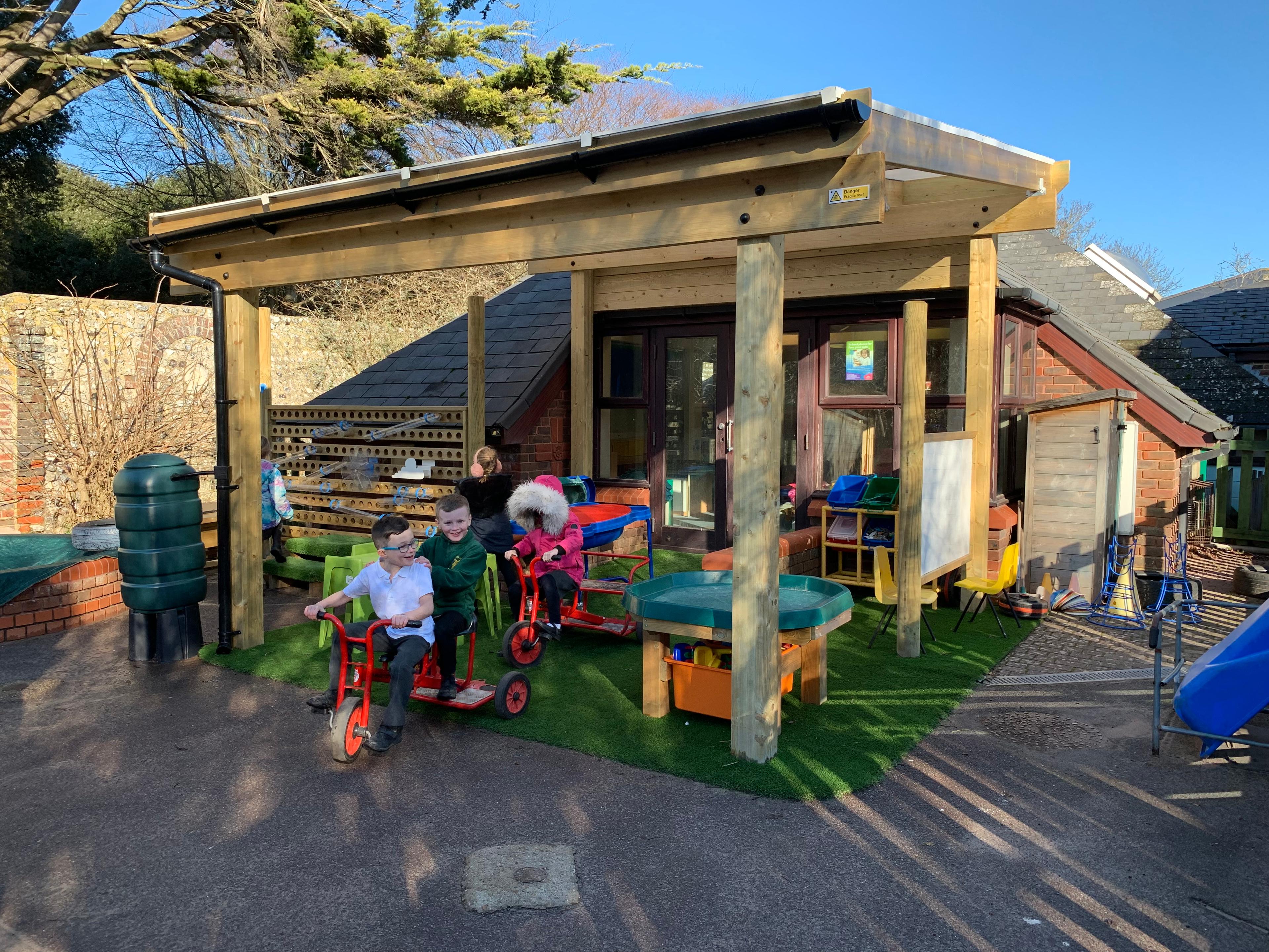 A Timber canopy that has been installed in an EYFS playground. An artificial grass surface has been placed in the canopy. A group of children are playing underneath the timber canopy with a wide variety of outdoor play equipment.