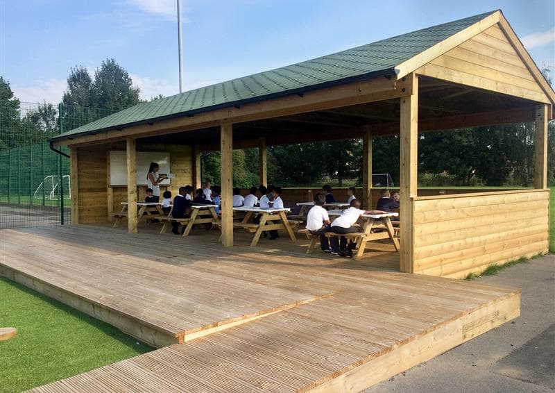 A wooden Gable-end outdoor classroom with picnic tables set up inside it. A whiteboard is set up at the front of the structure, with a teacher stood next to it and teaching a class.