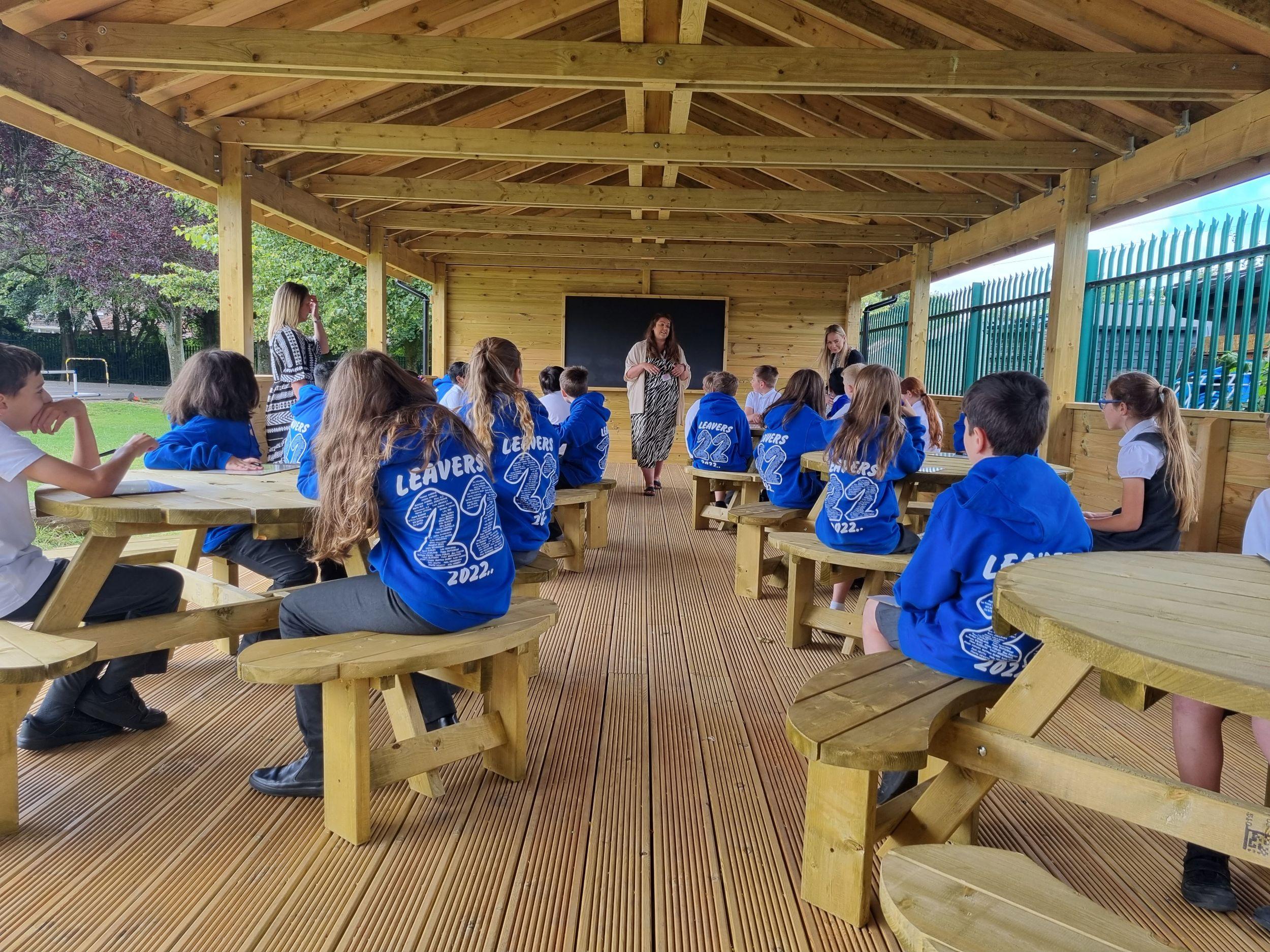 Students in blue leavers hoodies sit at circular picnic tables inside a large timber Gable-End outdoor classroom by Pentagon Play, while a teacher stands at the front by a chalkboard.