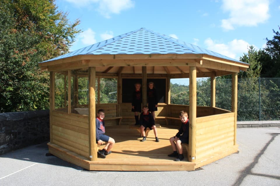 A wooden gazebo has been installed on a concrete surface. The gazebo is shaped like an octagon and has a blue roof. 5 children can be seen inside, looking at the camera and smiling.