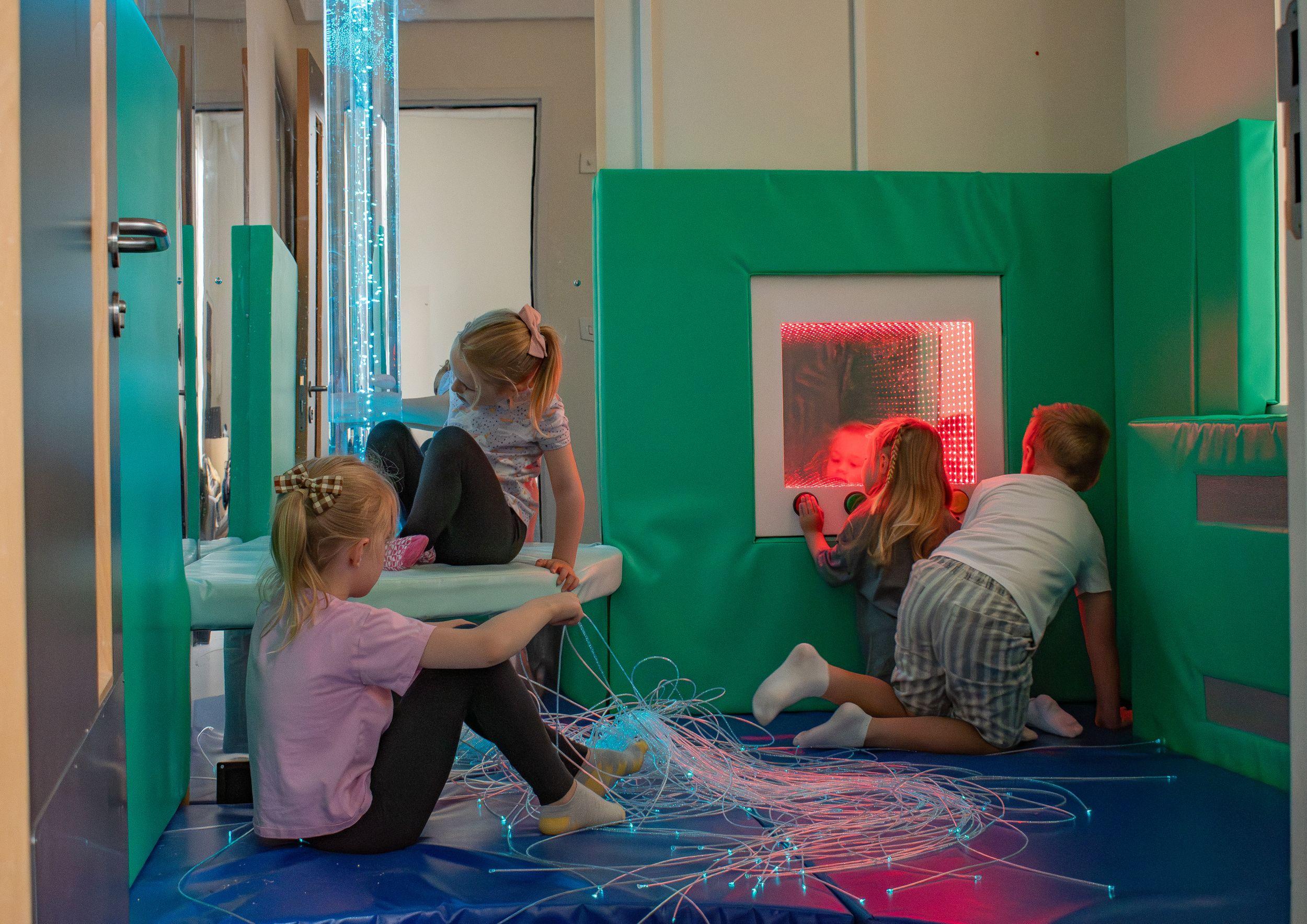 Children explore a sensory room featuring a glowing blue bubble tube, a red deluxe infinity panel, and a bundle of sparkling fibre optic sensory lights spread across a blue padded floor.