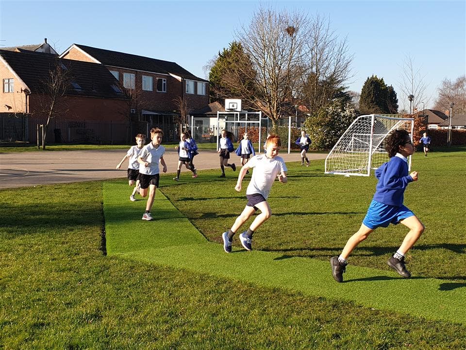 A class of children running on their daily mile track, in the middle of the daily mile track there is a goal with grass surrounding the track.