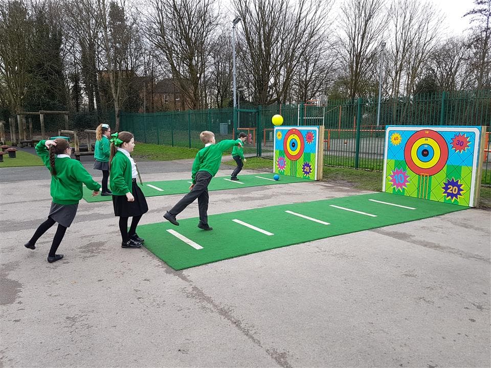 Two goal targets on a playground, with green Saferturf leading from the target with white marking lines. on the front goal target there are 3 children throwing a ball and on the furthest away target there are 2 children throwing a ball.