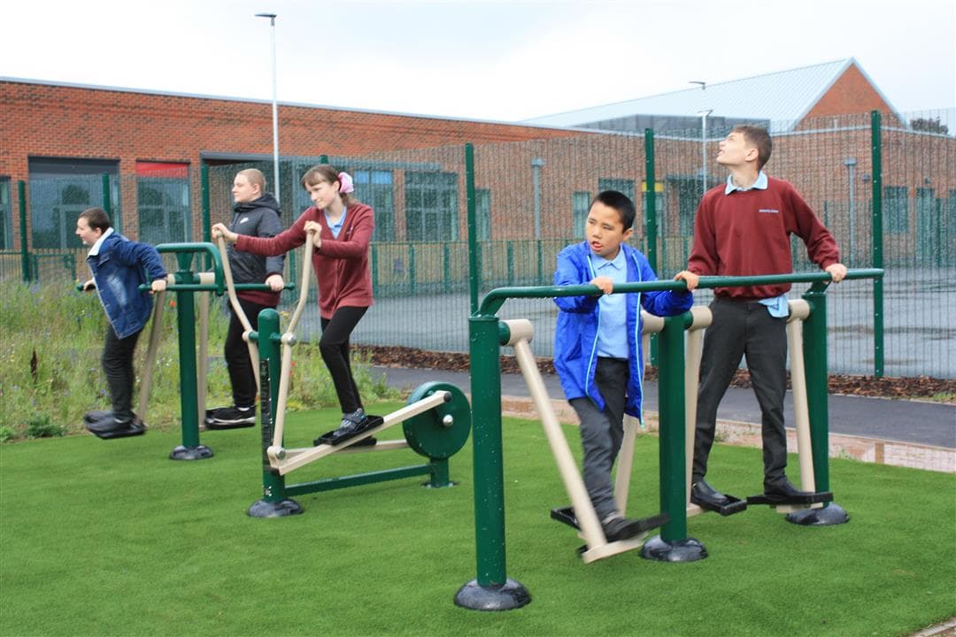 5 children playing on their outdoor gym, 2 children are on a double air walker and one girl is on a cross trainer, the other two boys are playing in the background.