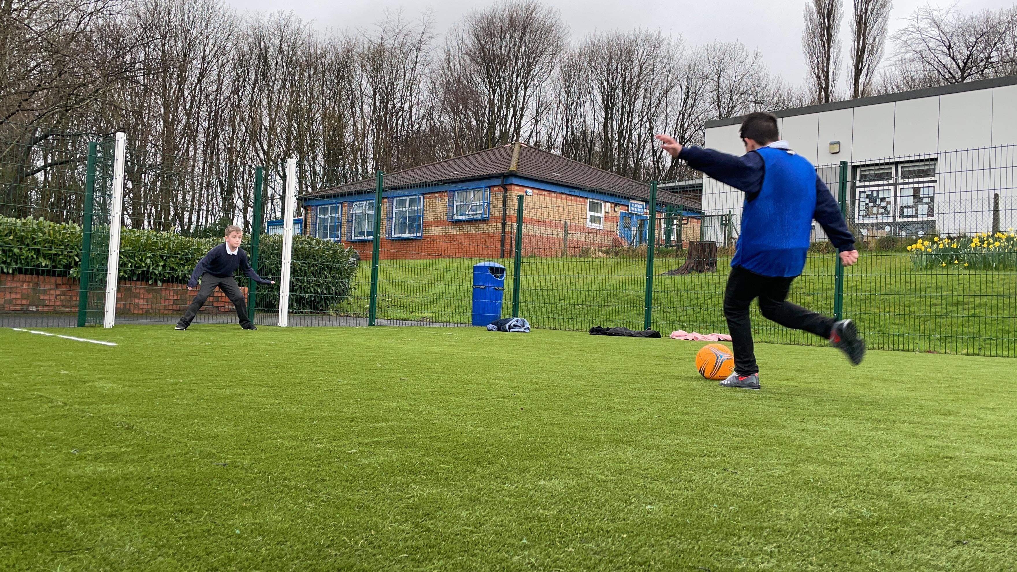 2 children playing on their multi-use games area, one boy is in net waiting to save the goal and the other young boy is taking a shot towards the goal
