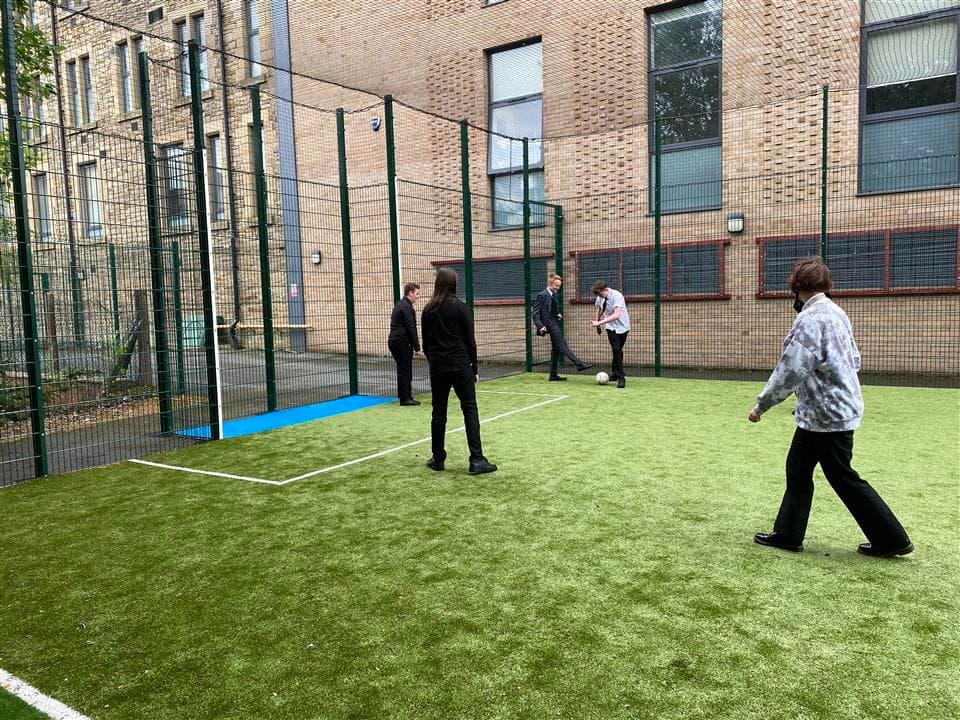 5 children playing on a recently installed MUGA at their school, one young boy is in the net and the rest are playing football.