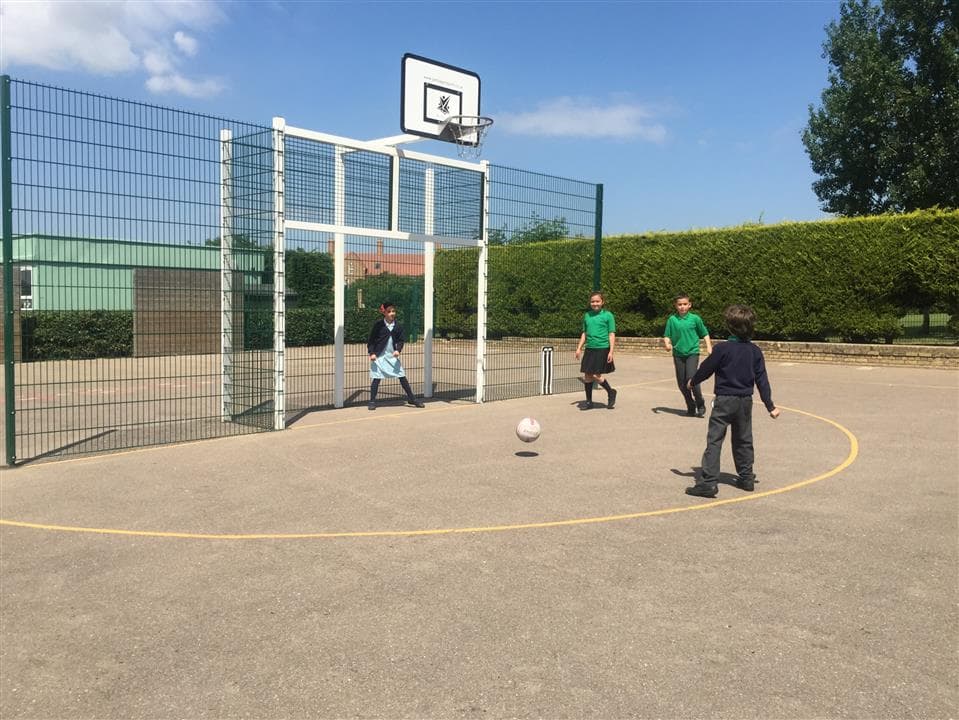 3 children playing football aiming at the maxi goal, one girl is stood within the goal posts waiting to save the ball.