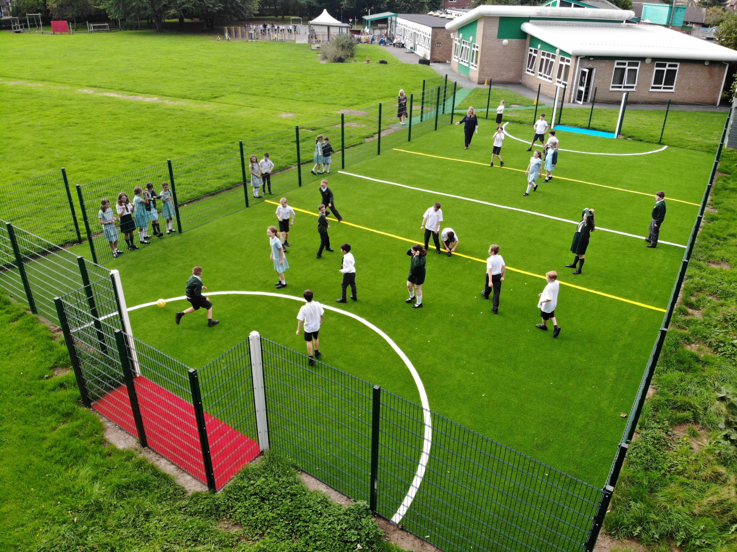 An aerial view of a Pentagon Play MUGA with green artificial grass and white sports markings, where school children in uniform are playing football within a fenced enclosure.