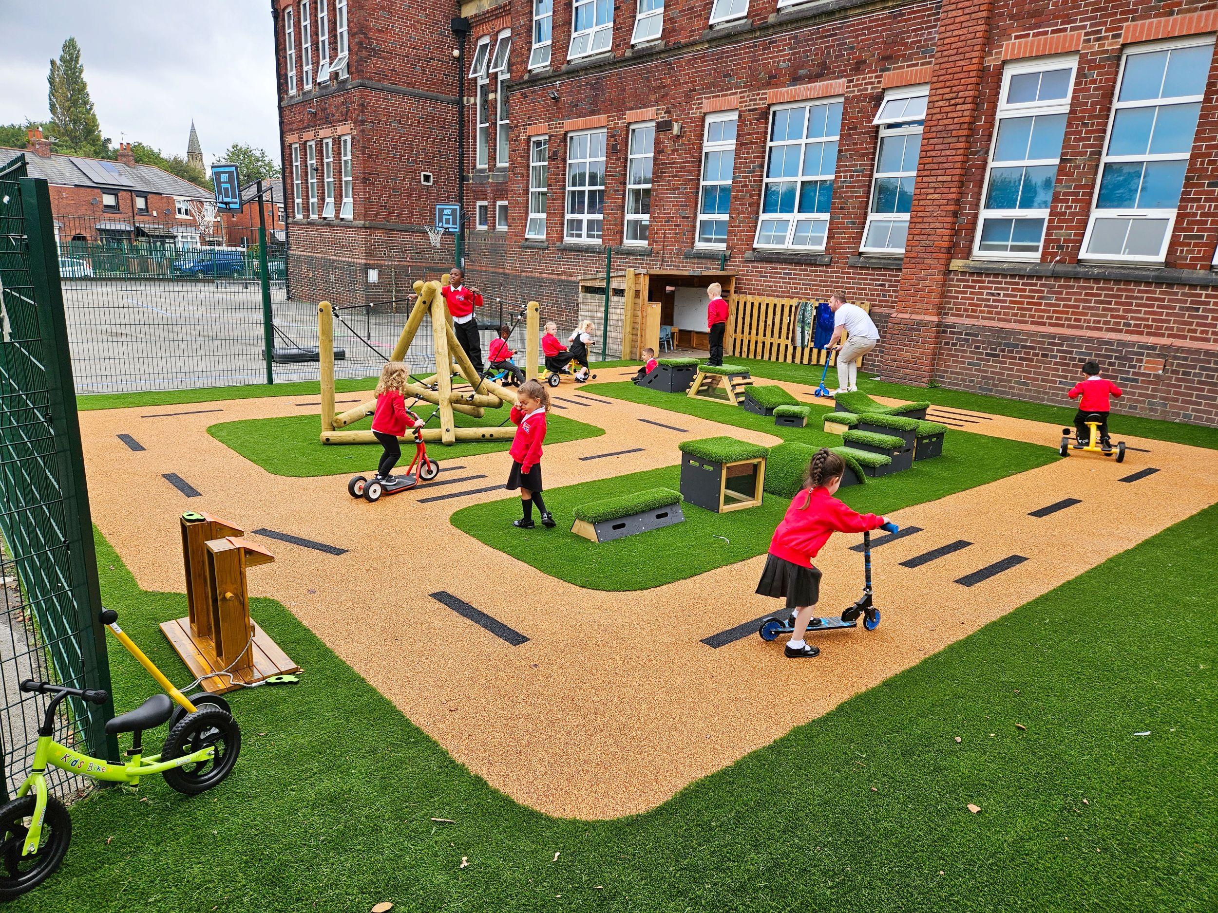 Children in red sweaters ride scooters and trikes on a bespoke wetpour roadway track, surrounded by green artificial grass and timber play equipment from Pentagon Play in a school playground.