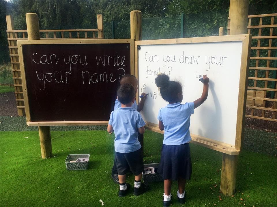 3 children drawing on a giant white board with the giant chalk board next to them. 2 of the children are writing and one of the children on watching.