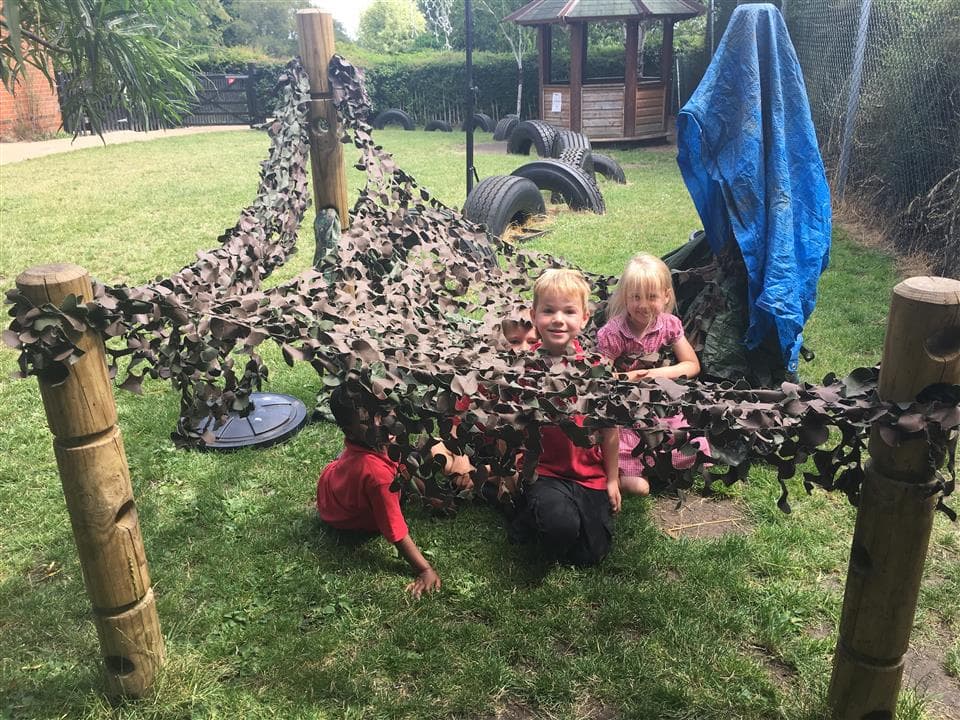 3 children playing underneath their den making posts, they have used a leaf net over the top to create a shelter.