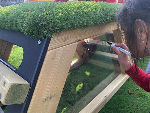 A school child drawing on clear panel on the Get Set, Go Block with a black marker. The top of the Get Set, Go! Block is artificial grass, with the clear panel being surrounded by natural wood.