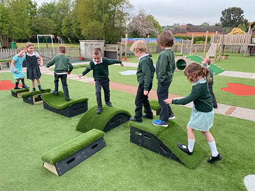 School children playing on Get Set, Go! Blocks, which have been placed on an artificial grass surface. The children are in a single file line as they traverse the obstacle course they have created.