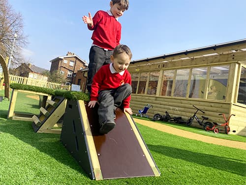 School children playing on Get Set Go Blocks, which have been placed on artificial grass. One child is sliding down a slide block whilst the other child is stood on the top of the block, looking down the slide.
