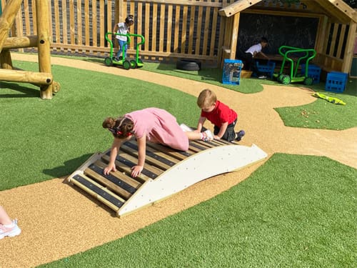 School and nursery children climbing on the Play Bridge. The play bridge has been placed on a wetpour path, with artificial grass surrounding the path. The children are crawling on behind the other over the bridge.