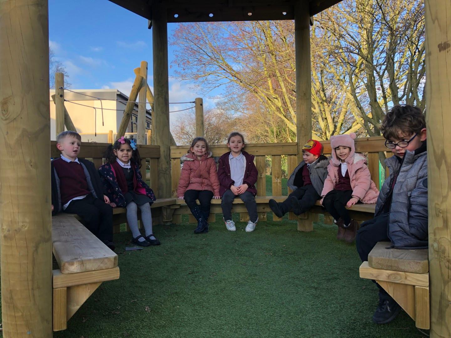 A group of children are sat on internal benches that are within a wooden gazebo. The children are all looking at the camera and smiling. Trees can be seen in the background, surrounding the kids in nature.