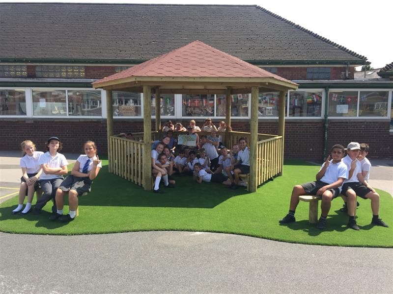 A wooden gazebo has been installed on artificial grass, with a bench installed on both the right and left side of it. A group of children are sat and stood up in the Gazebo, with 2 sets of 3 children sat on one of the benches outside the Gazebo.