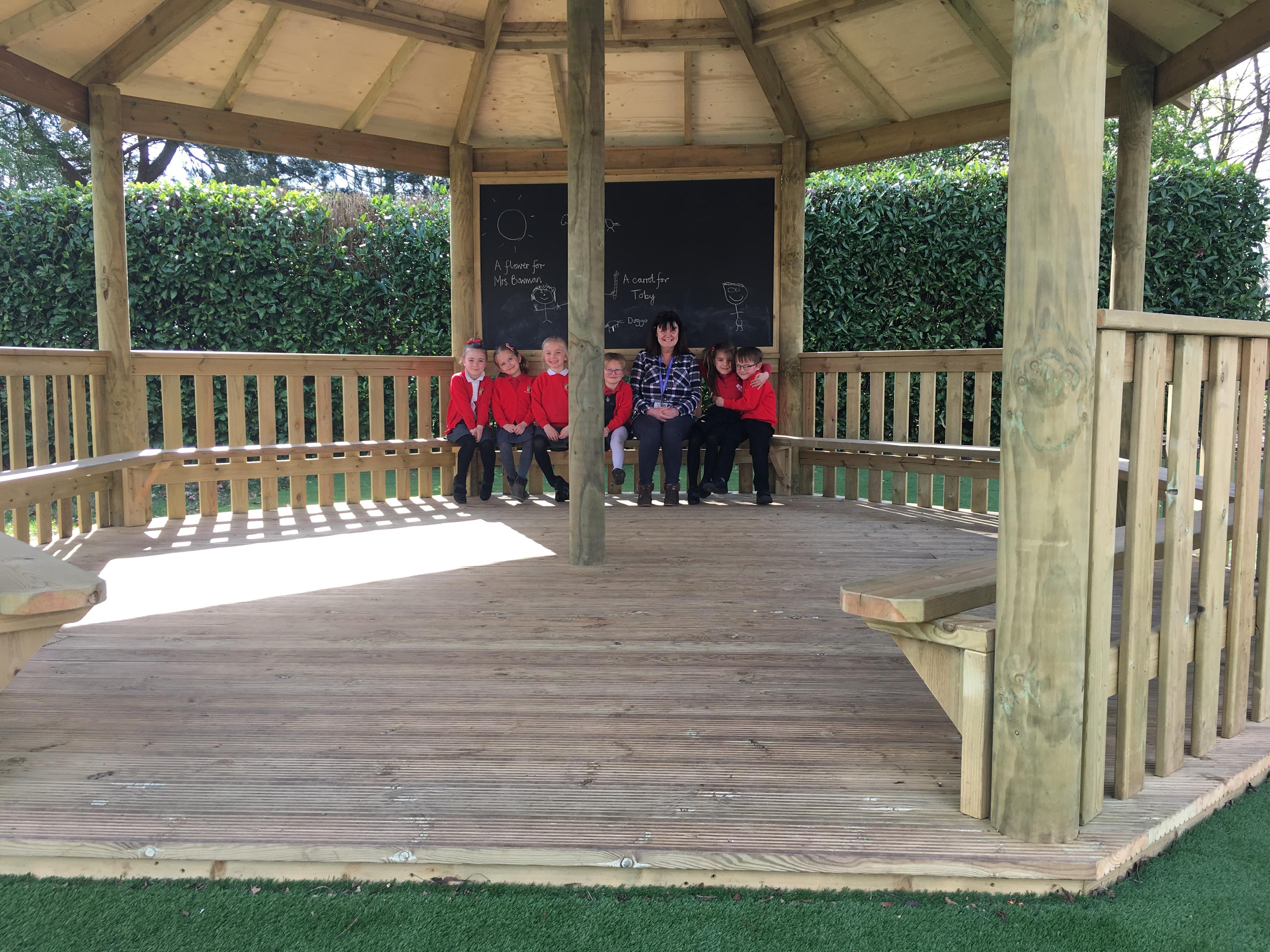 A group of children are sat inside a wooden gazebo, with a teacher sat in the middle. A wooden bench can be seen going around the entire gazebo. A clear view of the wooden flooring can be seen and a giant chalkboard can be seen behind the group of students and teacher.