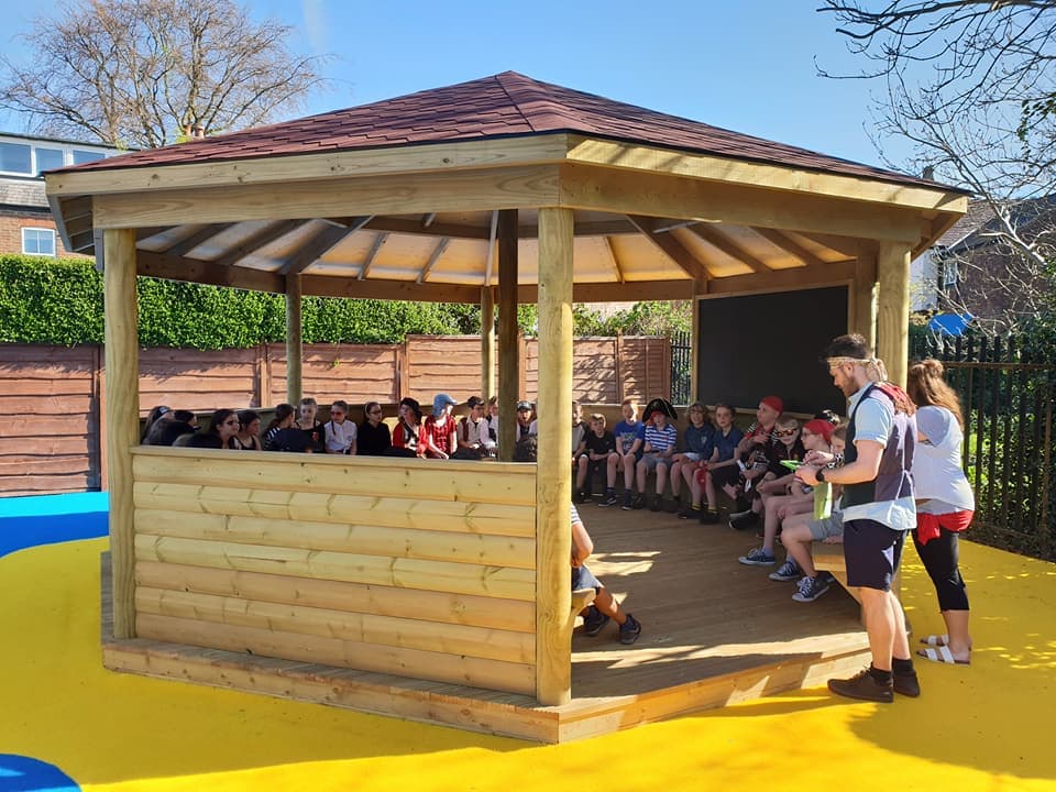 A wooden gazebo that has been installed on top of yellow wetpour. A huge group of children are sat all around the gazebo as two teachers stand at the entrance. The teachers are giving a lesson to the kids as they all look at the teachers.
