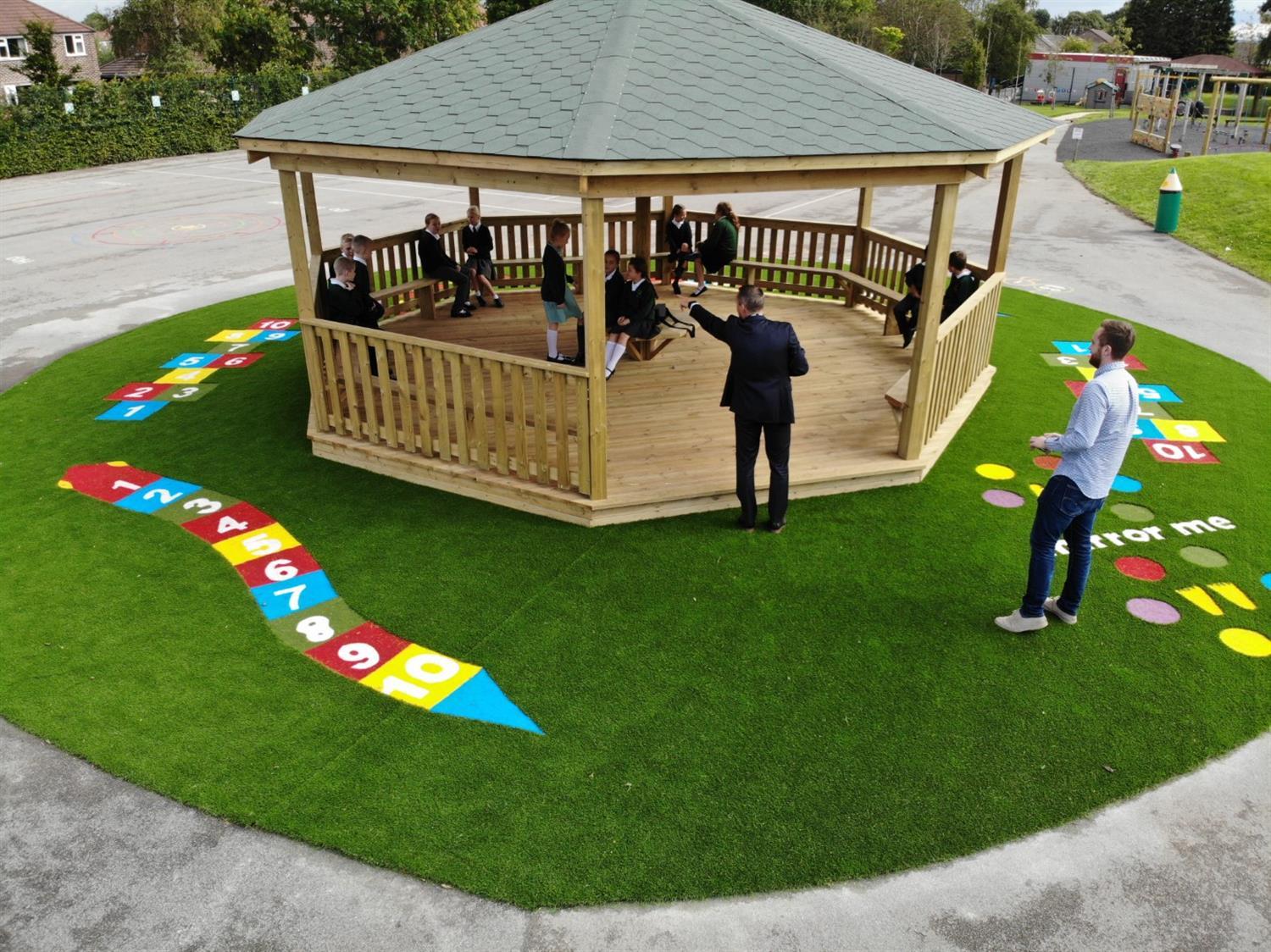 A wooden Gazebo that has been installed on an artificial grass surface and has Saferturf graphics around the Gazebo. The Gazebo currently has a group of children inside it and 2 teachers leading a lesson. Trees and natural grass fields can be seen in the background.