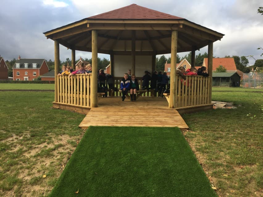 A wooden gazebo installed on natural grass, with a wooden ramp that connects the entrance of the structure to an artificial grass path. In the inside of the wooden gazebo, a group of children can be seen sat around and looking in different directions, taking in the nature that surrounds them.