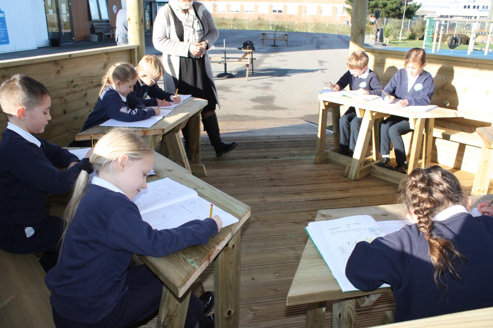 A group of children are working at desks that have been set up within a wooden gazebo. The children are writing in their books as a teacher watches over them. The playground can be seen in the background, being a tarmac playground.