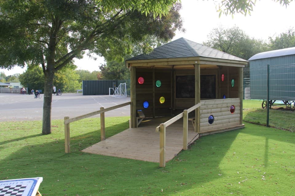 A wooden gazebo with a wooden ramp leading to the entrance of the structure. A variety of circular windows can be seen installed on the side of the Gazebo, with coloured plastic covering the holes. The colours are red, green, blue and yellow.