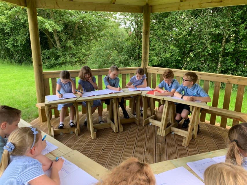 A wooden gazebo with a lot of children inside. The children are sat at desks and are writing on paper. The children are surrounded by trees and grass fields, providing a sense of nature.