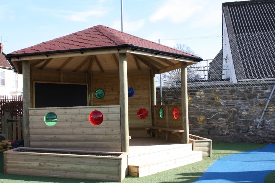Wooden gazebo that has been installed on green wetpour. The walls of the gazebo have circles cut out of them, with them being coloured red or green. The roof of the structure is red, with planters being placed on the left and right of the entrance to the Gazebo.
