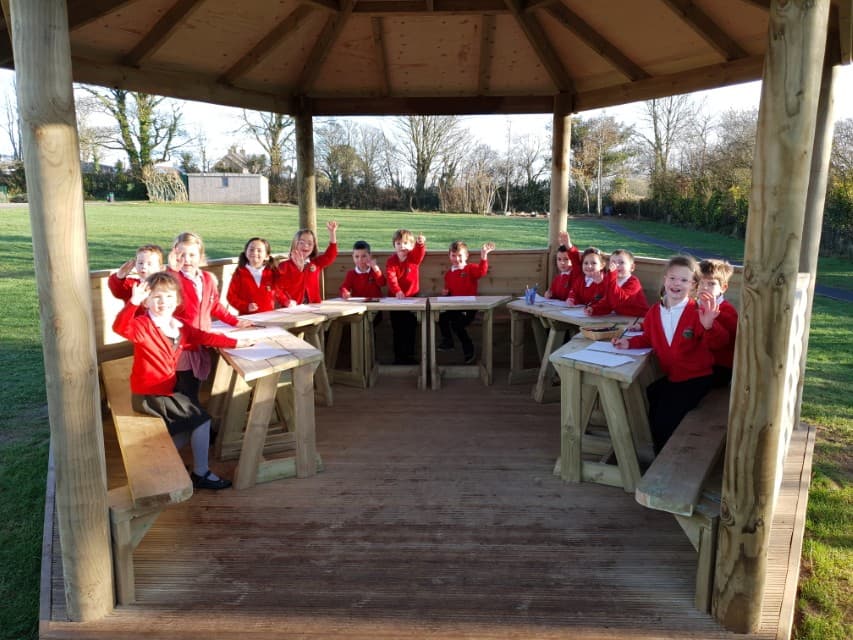 A group of children are sat on benches that are attached to the inside of a wooden gazebo. They all have small tables in front of them with work placed on them. The children are all looking at the camera and smiling. In the background, you can see trees and grass, creating a natural environment.