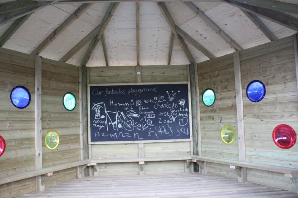 The inside of a wooden gazebo. The back wall has a chalkboard attached to it, with the rest of the walls have 2 circles cut out on each panel. Over each circle is a coloured piece of plastic, such as red, blue, yellow, green.