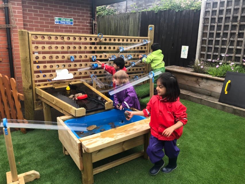 A group of children are playing and engaging with a variety of water play equipment. A water table, water dam and a water wall can be seen on the photo, all connecting to each other. The equipment is installed on an artificial grass surface.