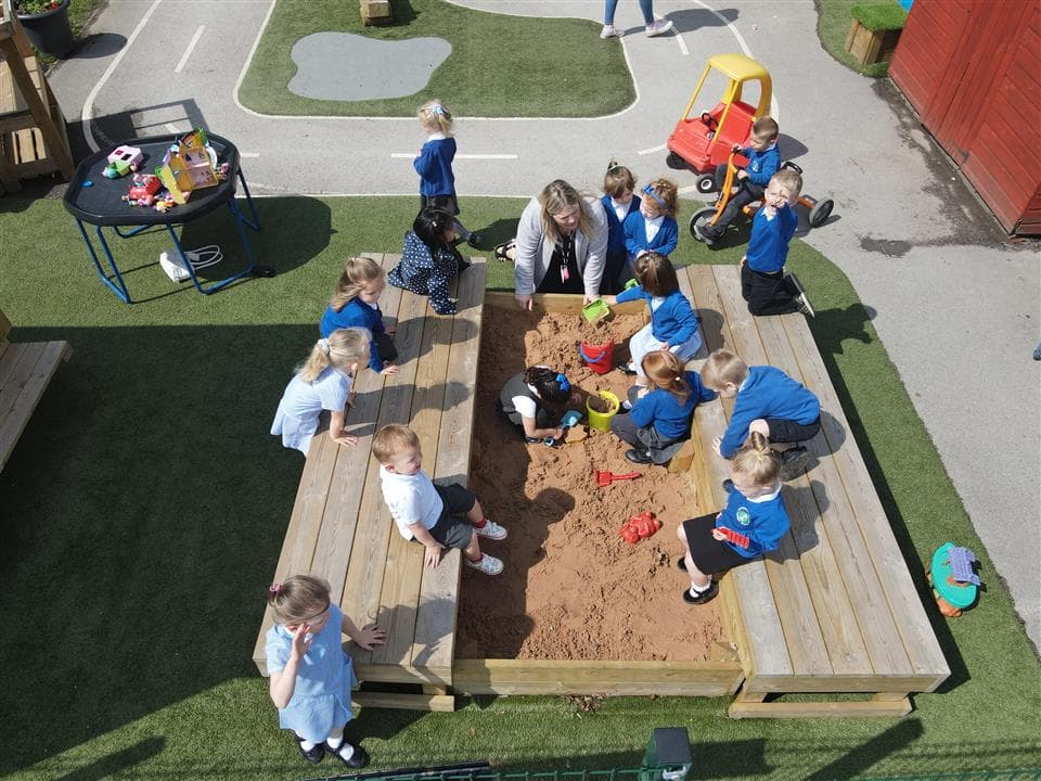 A class of children and a teacher are all playing in the sand box, they are digging and making sand castles. 