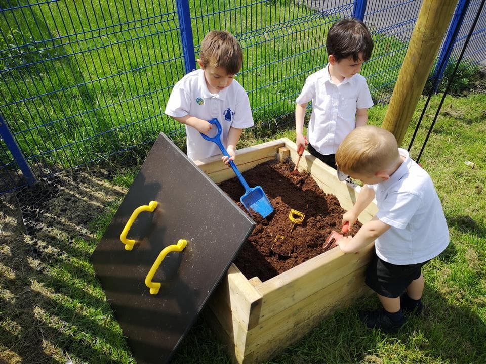 3 children playing in their mud box, there are spades and toys within the box. The Mud Box has been placed on a natural grass turf, with the boys looking in the box.