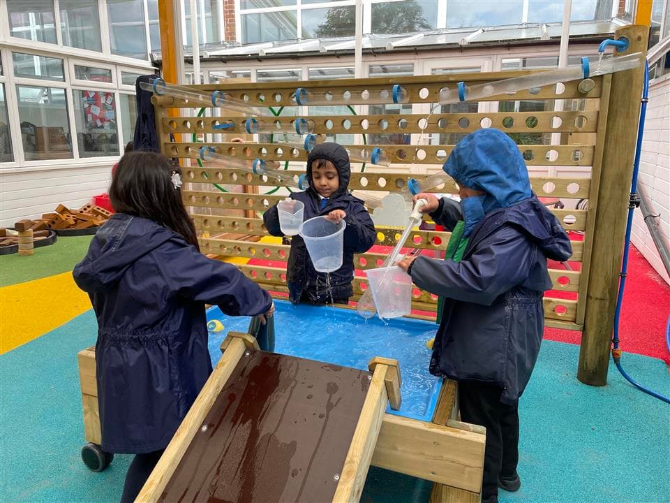 3 children playing on the fine motor table, they are all wearing rain coats and using jugs to fill water and pour down the damming station. 