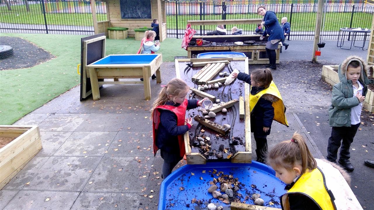 3 children playing on their damming station, they are using bottles of water to do their experiment, there is rocks and pieces of wood to act as the dam
