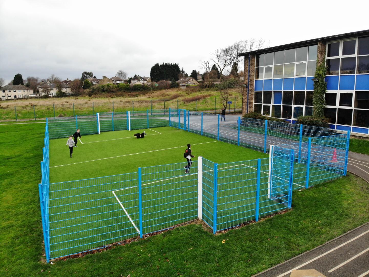 An artificial grass MUGA with a light-blue fence going around it. The court has built in football goals, with white posts. There are white and yellow line makings on the artificial grass.
