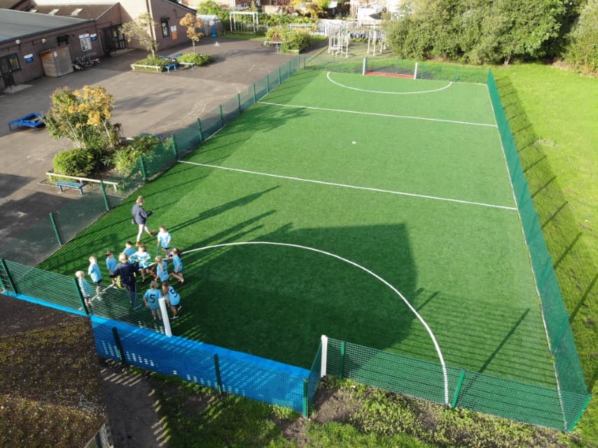 A green artificial grass MUGA surface, with a green fence going around the entire pitch. A group of kids can be seen on one side of the pitch, grouping around the goal. The goals have white posts and have either a blue or red wetpour surface.