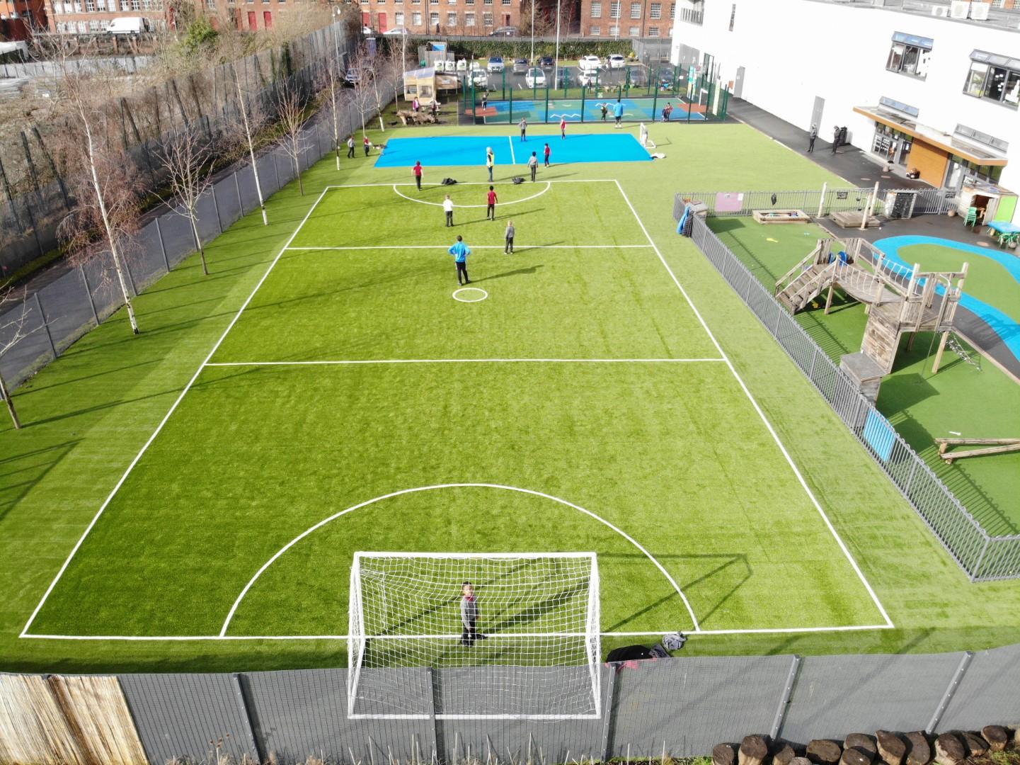A MUGA pitch that has been installed right next to a playground. The pitch has football line markings, with a group of children playing a match. The MUGA is in an open space with no fencing, with the school being seen in the background.