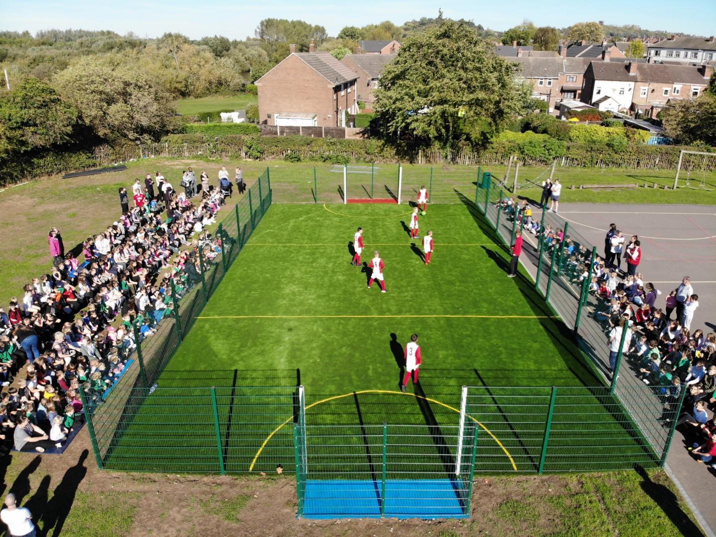 A Multi use games area with a group of children playing football on it. On both sides of the MUGA, a crowd can be seen watching the football match. The MUGA has been installed on a grass surface.