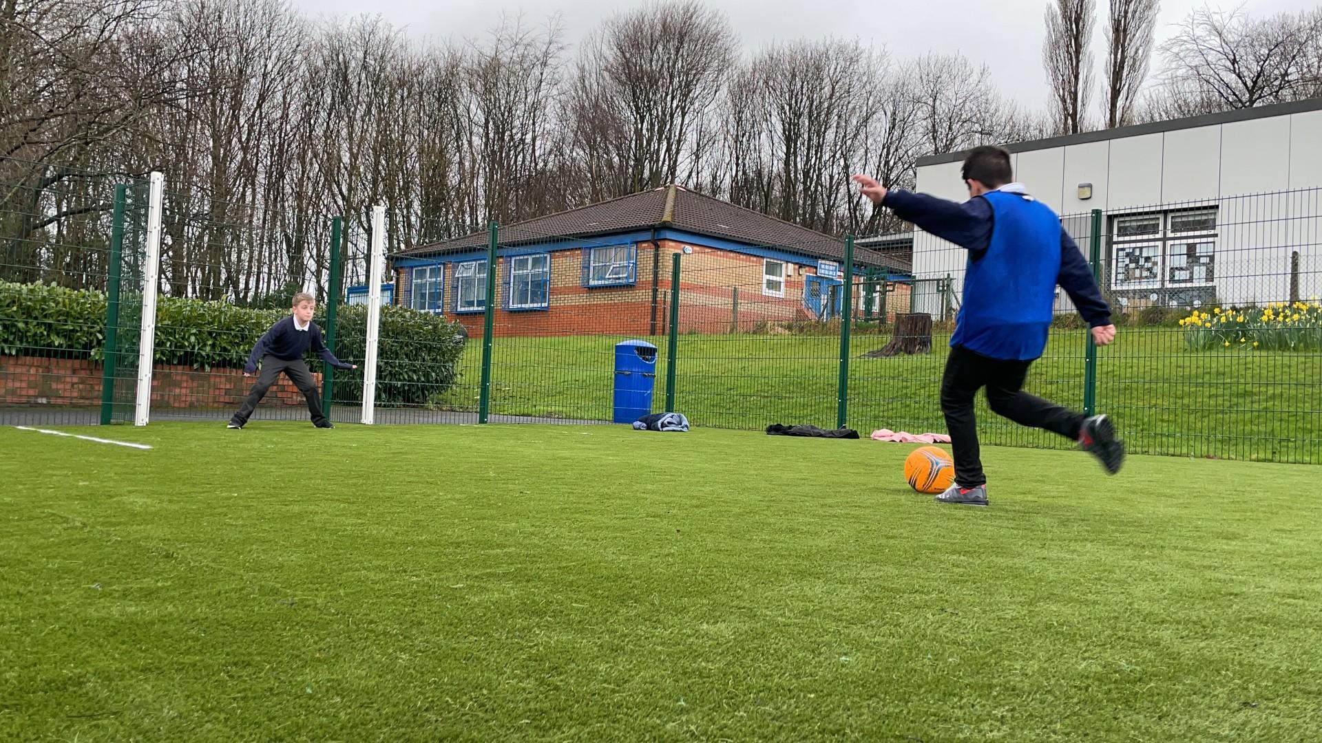 A Multi Use Games Area with an artificial grass surface. A child is taking a penalty kick whilst another child stands in goal. A green fence goes around the court, with the football goal's posts being coloured white.