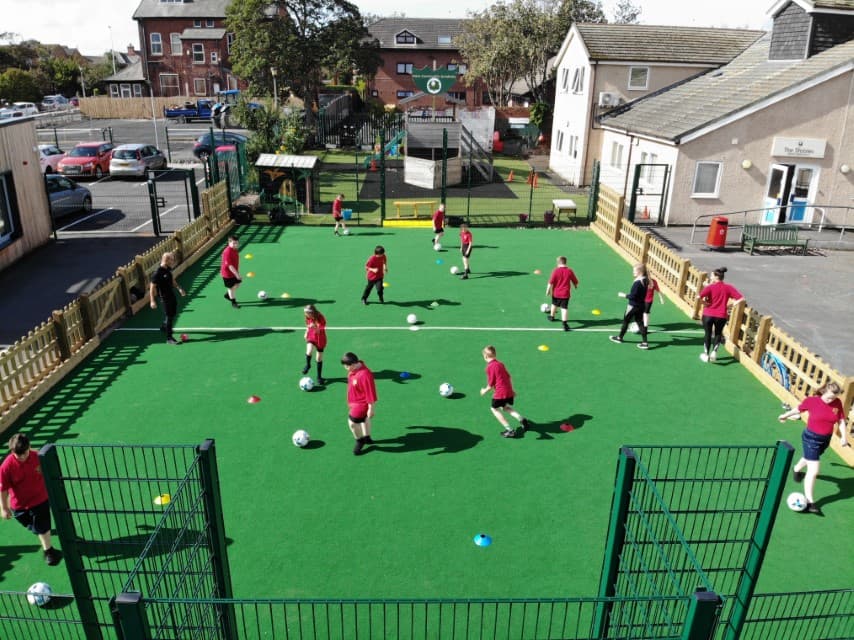 A MUGA Pitch with a dark green colour and a mixture of wooden and metal fencing. A group of children are playing with footballs on the pitch as they dribble the ball around cones. The goals are built into the metal fencing.