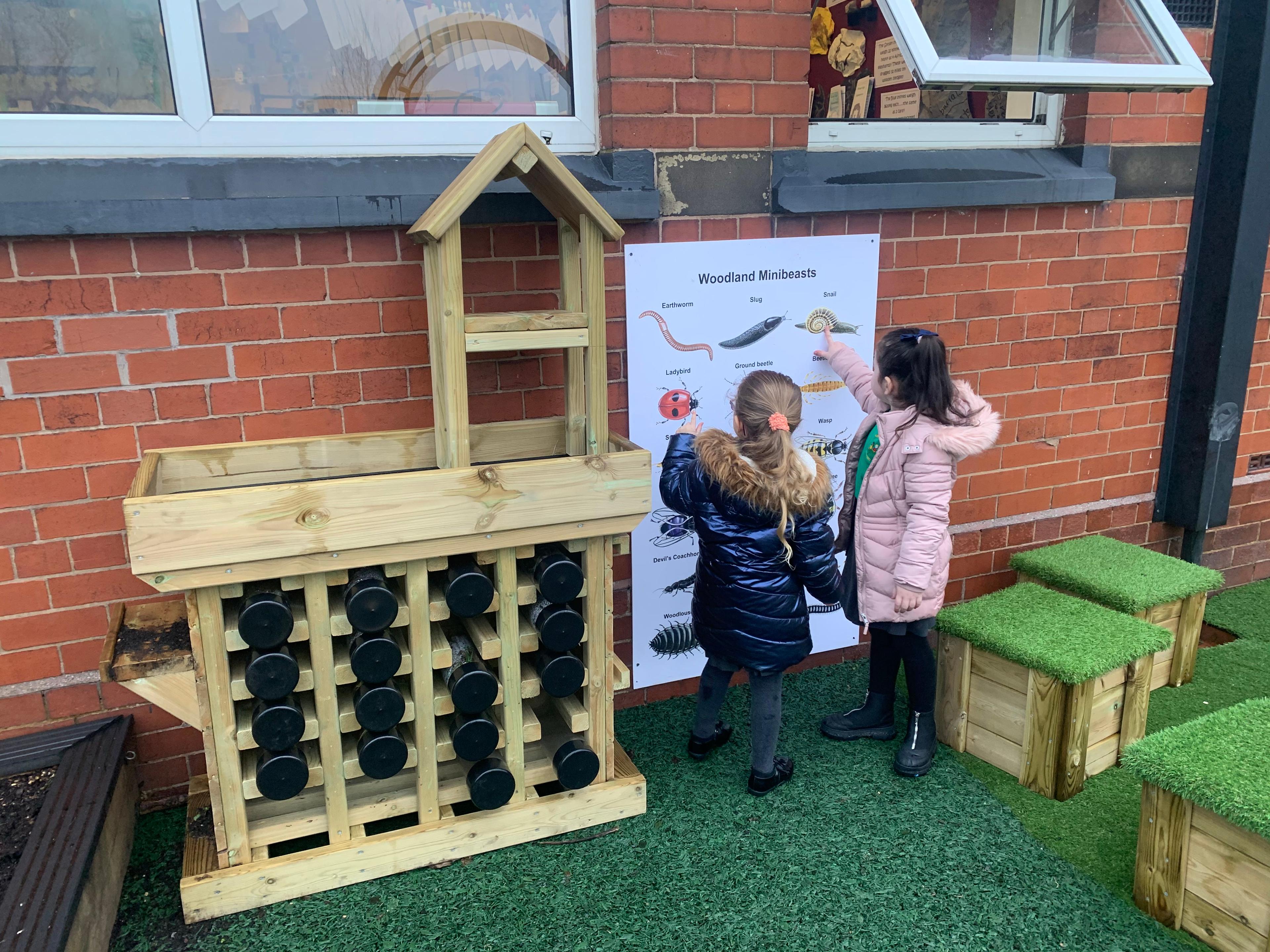 2 girls looking at their identifying bug board next to their bug hotel, there are grass top seats scattered around.