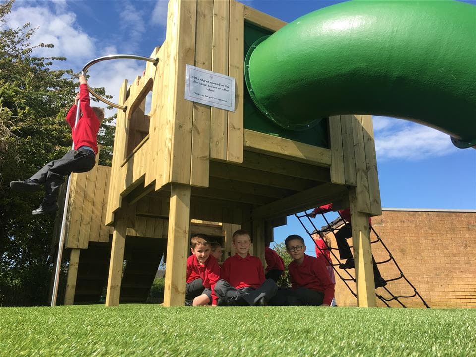 3 children playing around the langley play castle, 2 children are sat underneath and 1 child is swinging from the fireman's pole