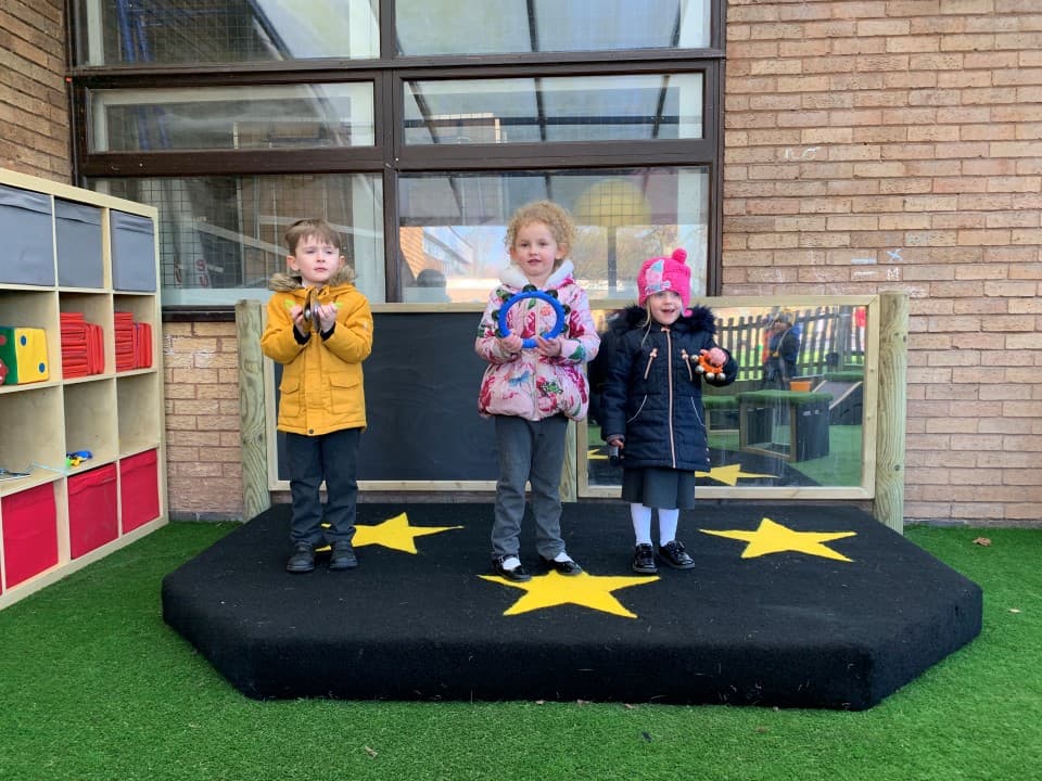 three children in winter coats and hats pose on the black performance stage with felt stars on the surfacing and a giant mirror and chalkboard in the back